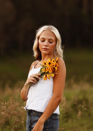 Young blonde woman holding yellow black-eyed Susan flowers with eyes closed in golden field at sunset.