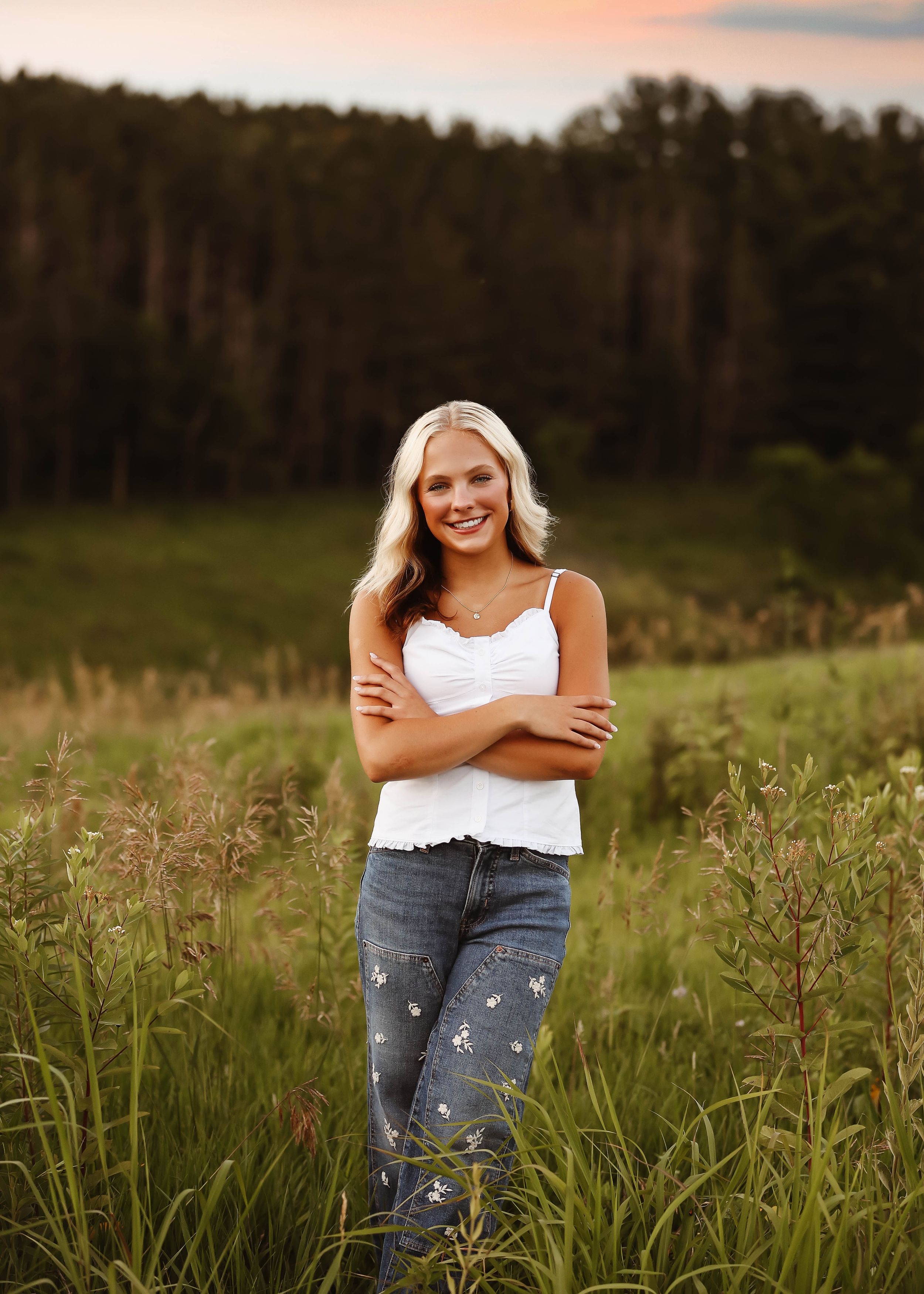 Smiling blonde teen poses with crossed arms in a lush green field at sunset wearing white top and floral jeans.