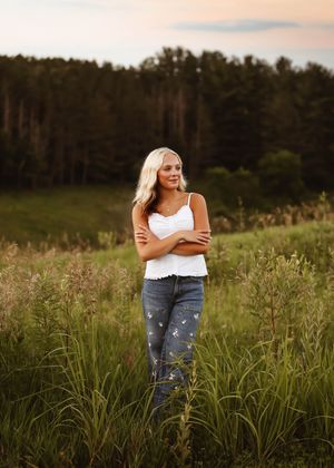 Blonde woman in white tank top and jeans stands in a lush green field at golden hour with forest backdrop.
