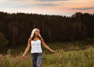 Blonde woman in white top and jeans stands in a meadow at sunset with arms outstretched, forest in background.