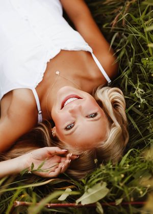 Smiling blonde woman lying in green grass wearing a white dress and delicate necklace, looking up joyfully outdoors.