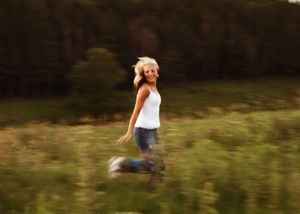 Joyful young woman in white tank top and jeans running through a lush green meadow with blurred motion effect.