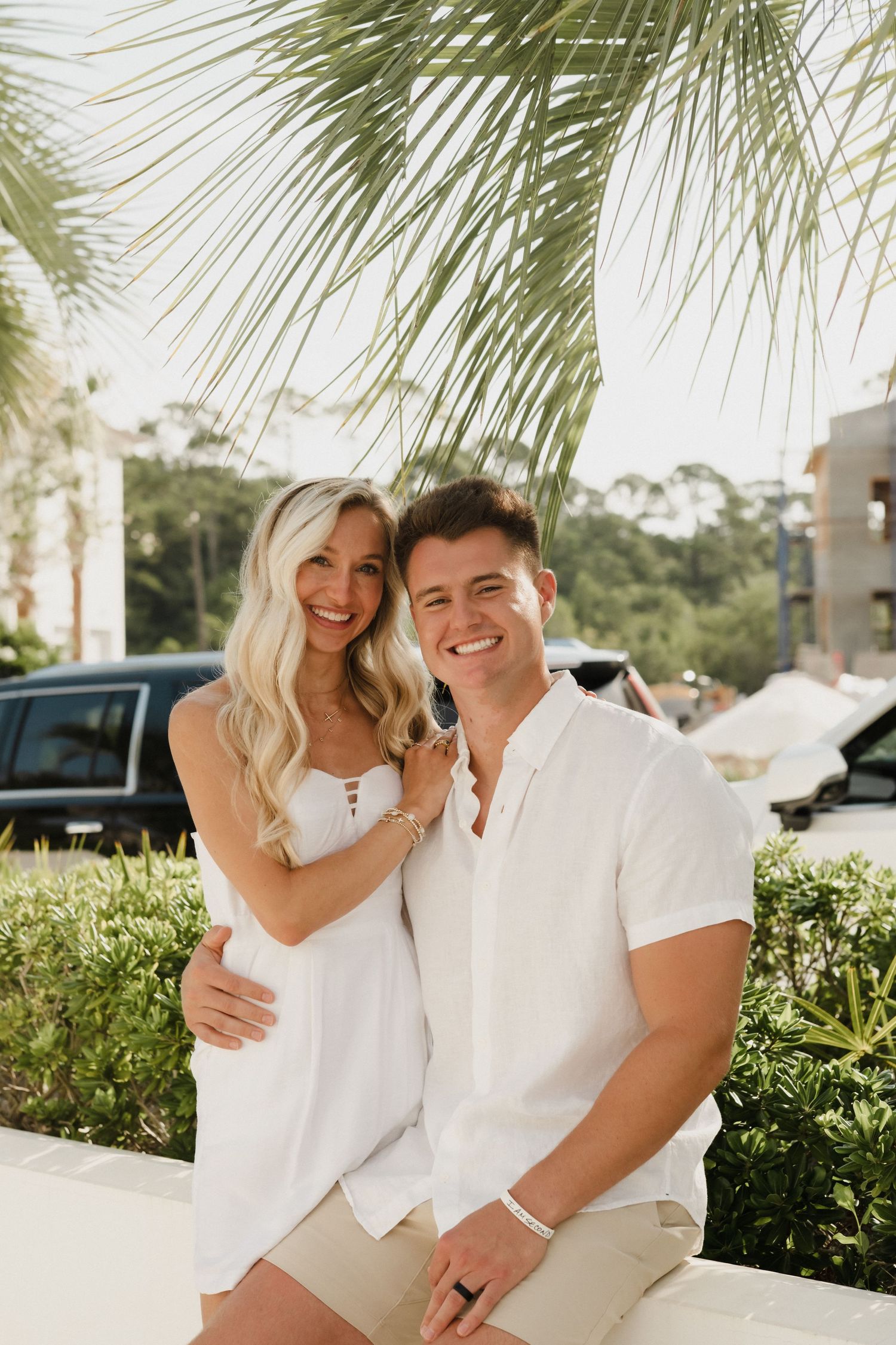 Couple wearing matching white outfits embrace and smile near tropical palm trees.