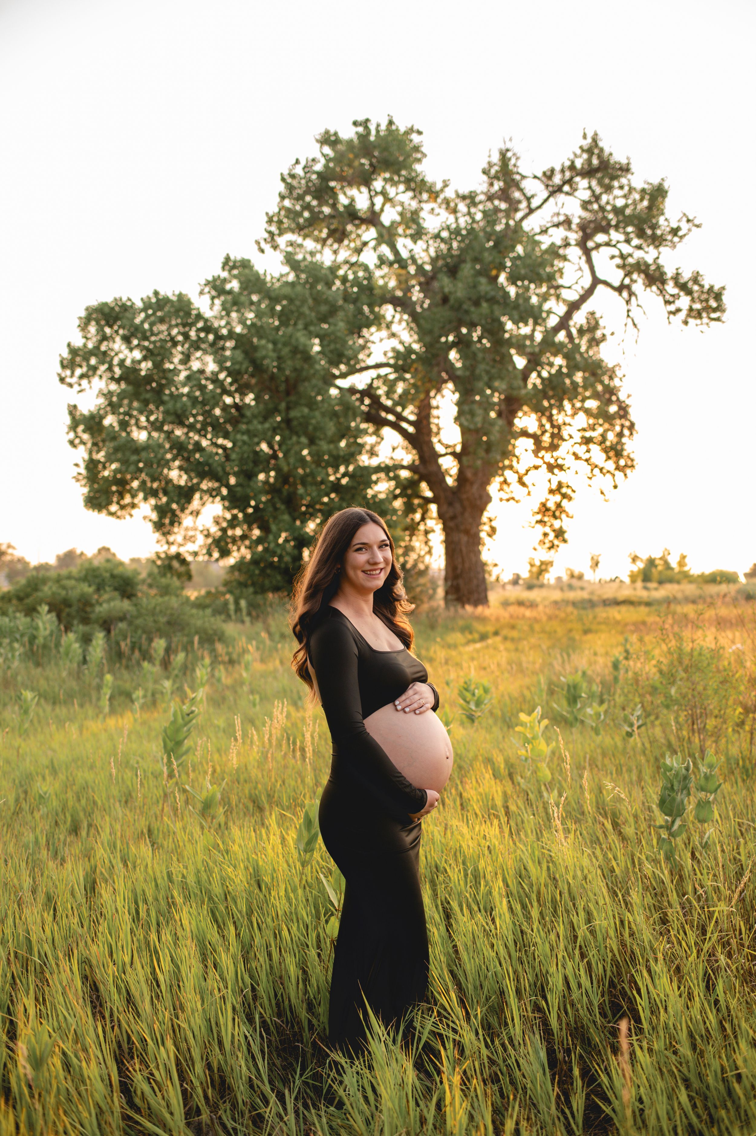Woman in black dress standing in golden sunlit field with large tree in background.