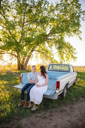 A couple sits together on a white pickup truck tailgate at sunset surrounded by grass and trees in a romantic rural setting.