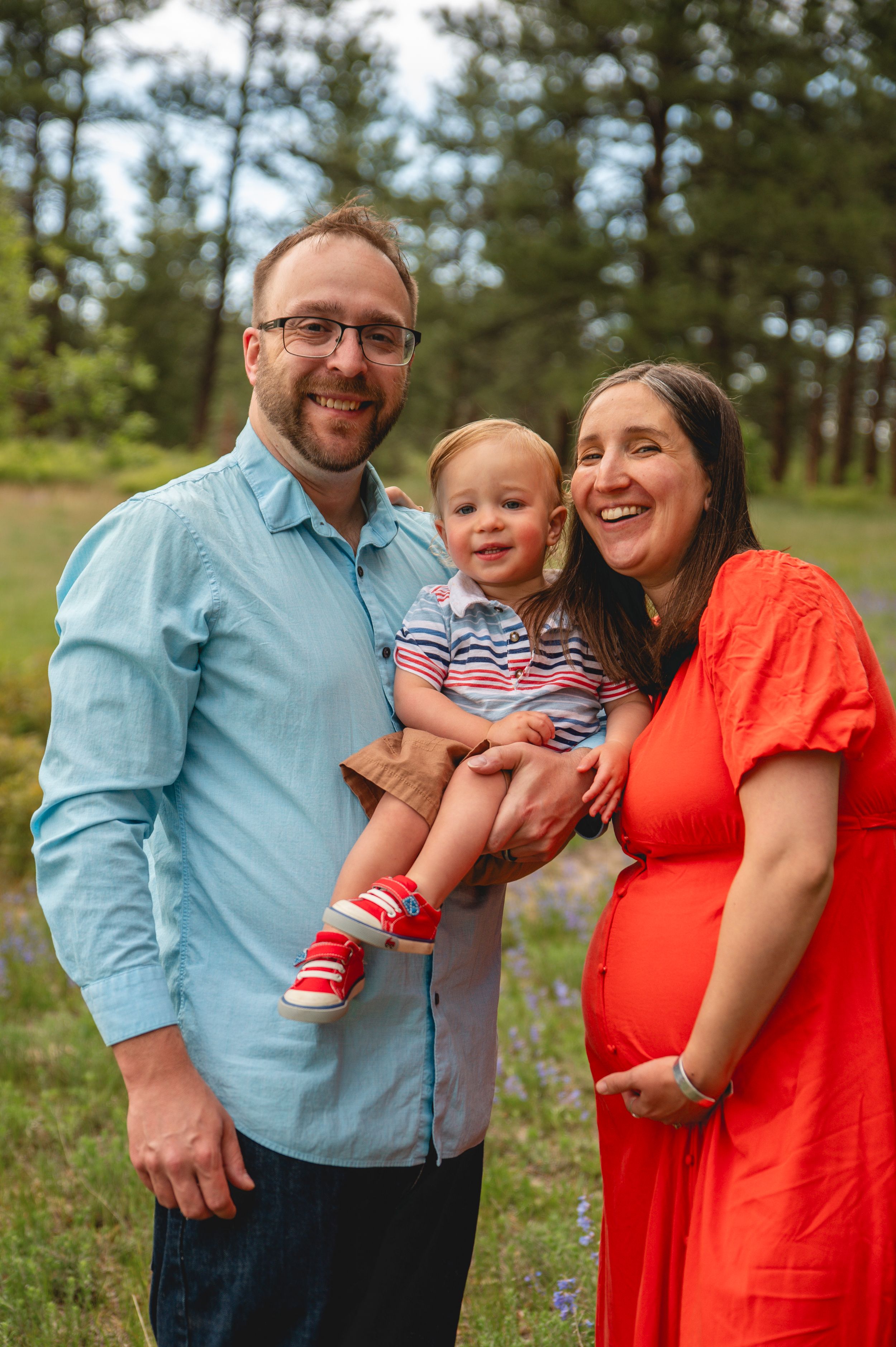 A family poses together outdoors in a wooded area with pine trees and wildflowers in the background.