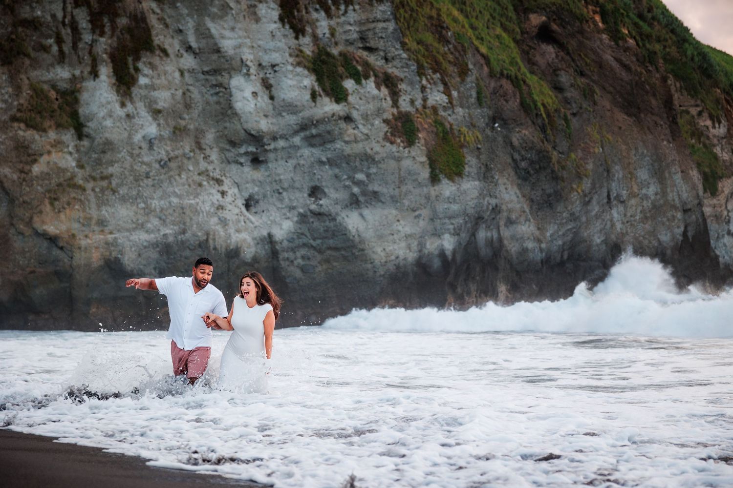 Couple laughing together as the ocean breeze blows along the Atlantic coastline, and a wave gets to them.
