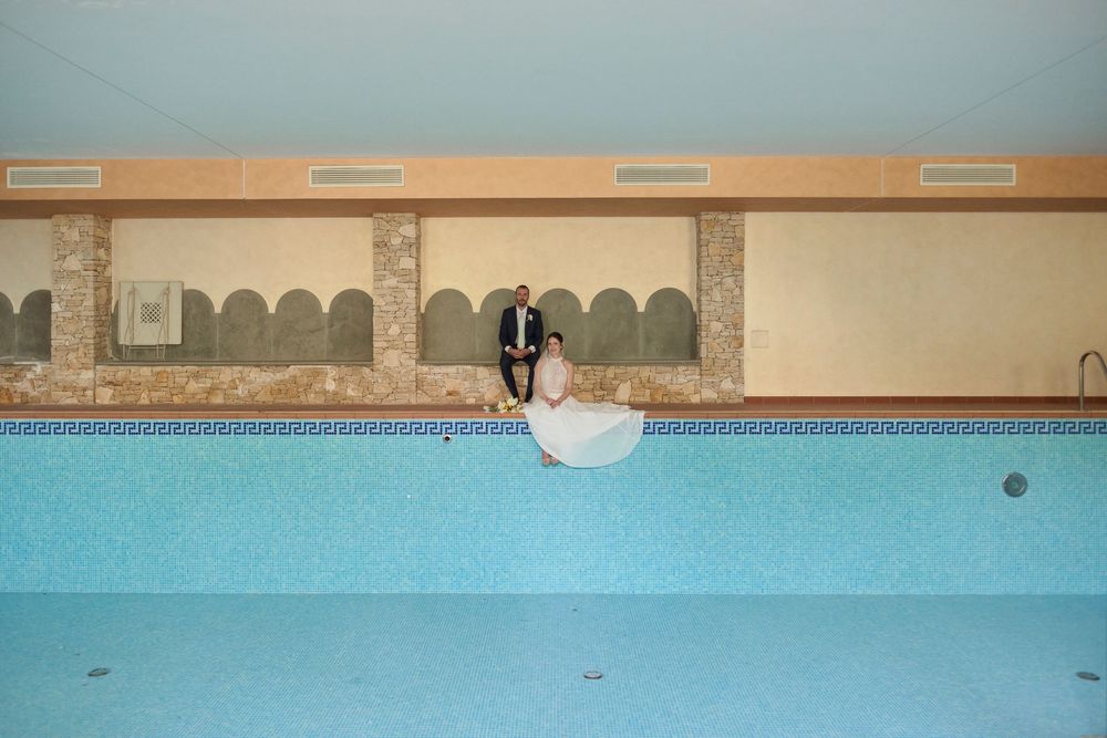 Bride in a white gown and groom in a dark suit sit together at the edge of an empty indoor swimming pool with arched stone walls.