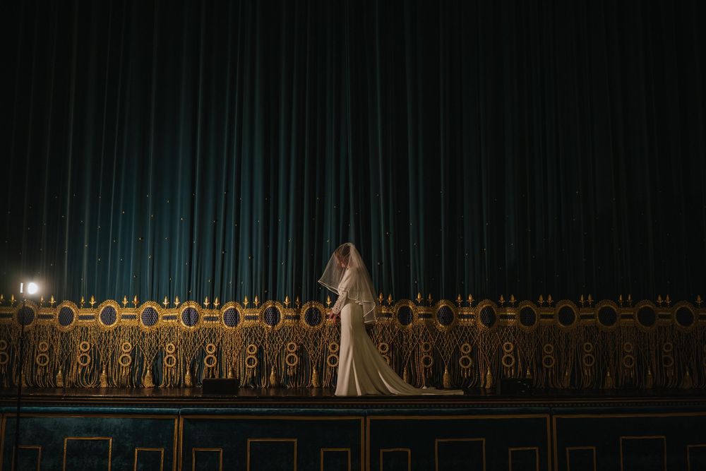 A bride in a white gown stands alone in a dimly lit cathedral with ornate wooden choir stalls behind her.