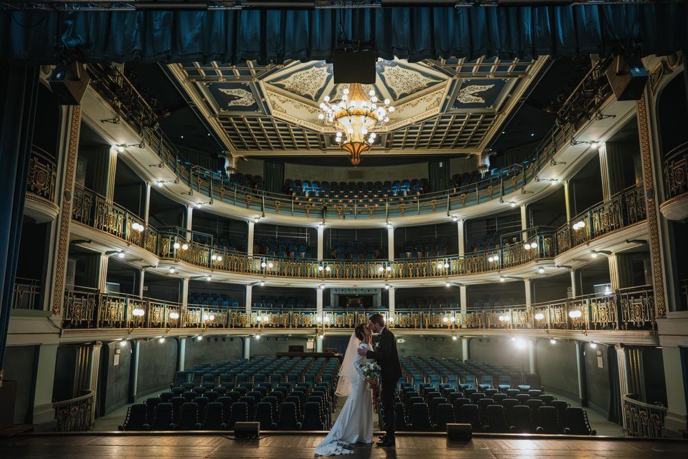 A couple in wedding attire embraces on stage in an ornate historic theater with tiered balconies and crystal chandelier.
