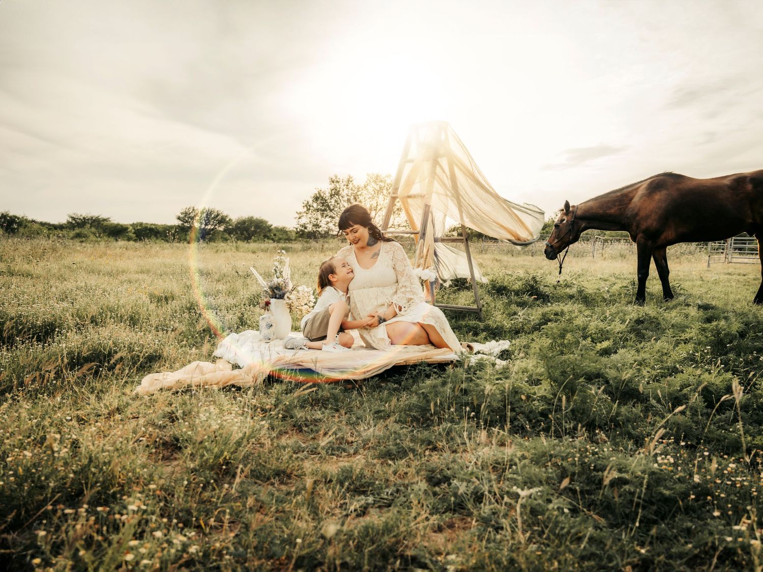 Mother and young son sitting on a blanket in a field, with a horse in the background, enjoying a peaceful moment together.
