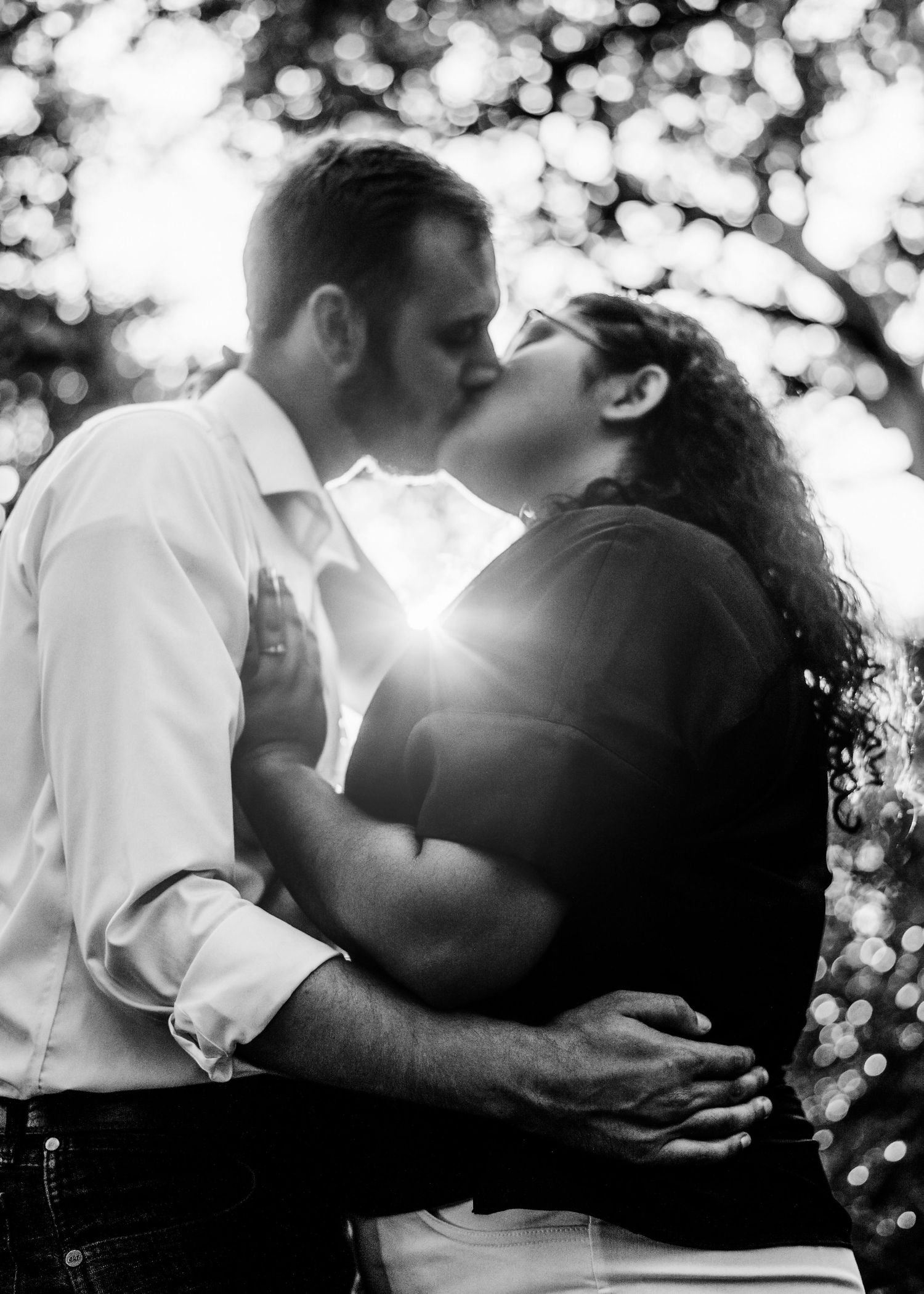 Black and white photo of a couple kissing, with dreamy light filtering through the tree, capturing a romantic moment.