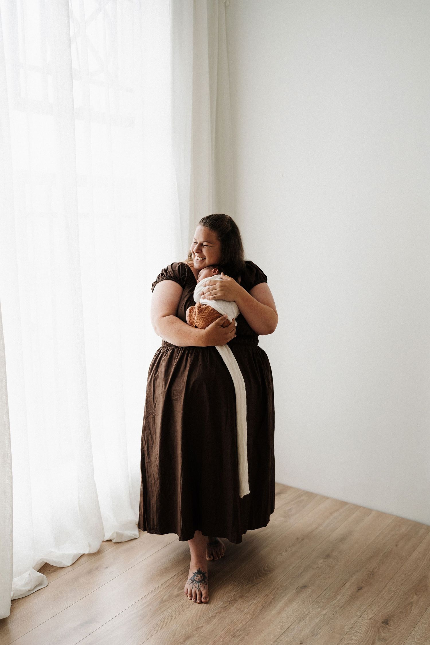 Parent in white sweater tenderly embraces sleeping newborn wrapped in beige blanket against neutral background.