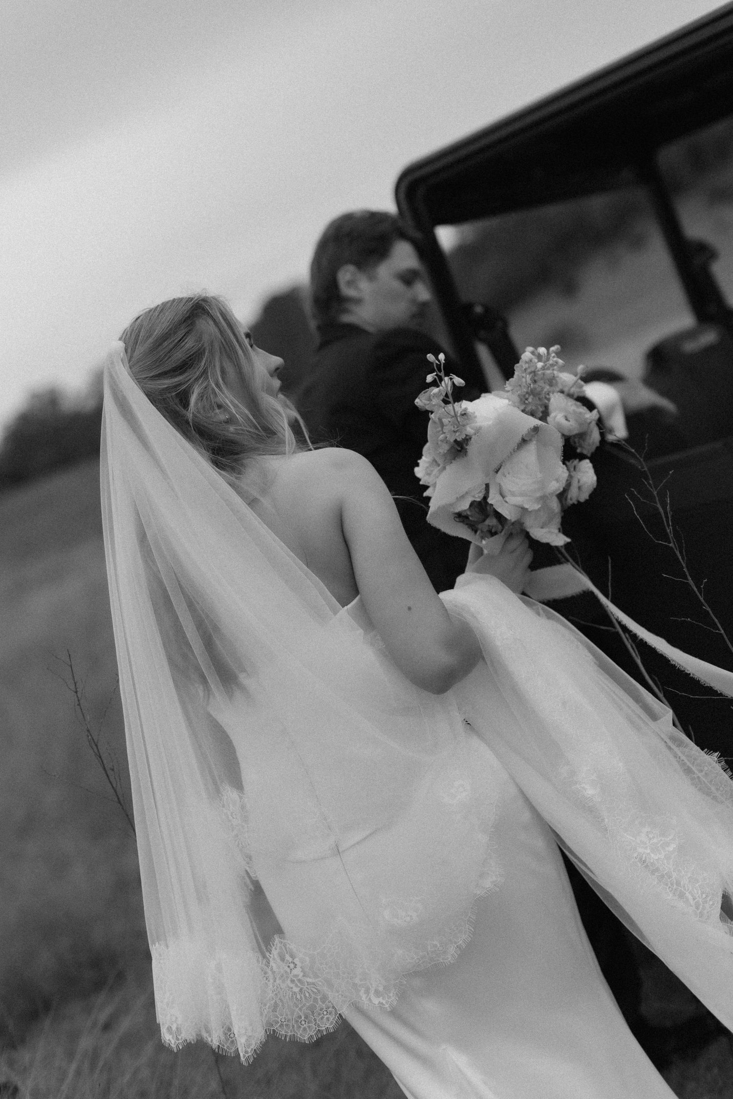 Newlyweds playfully posing with outstretched arms in a grassy hilltop field.