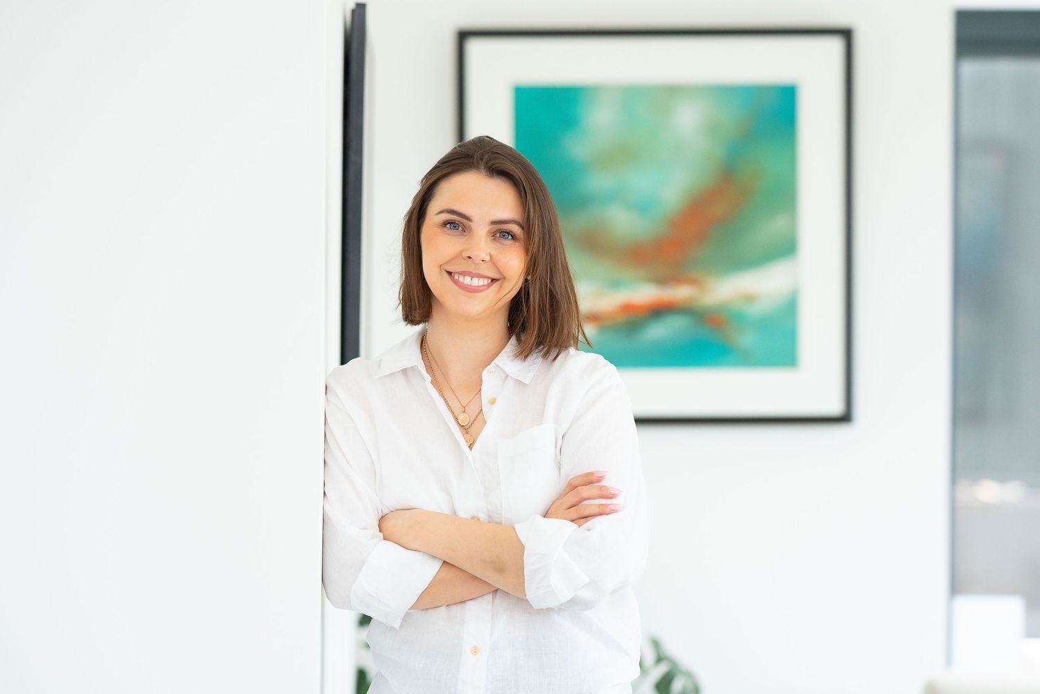 Professional in white blouse stands with arms crossed smiling in modern office with framed art on wall.