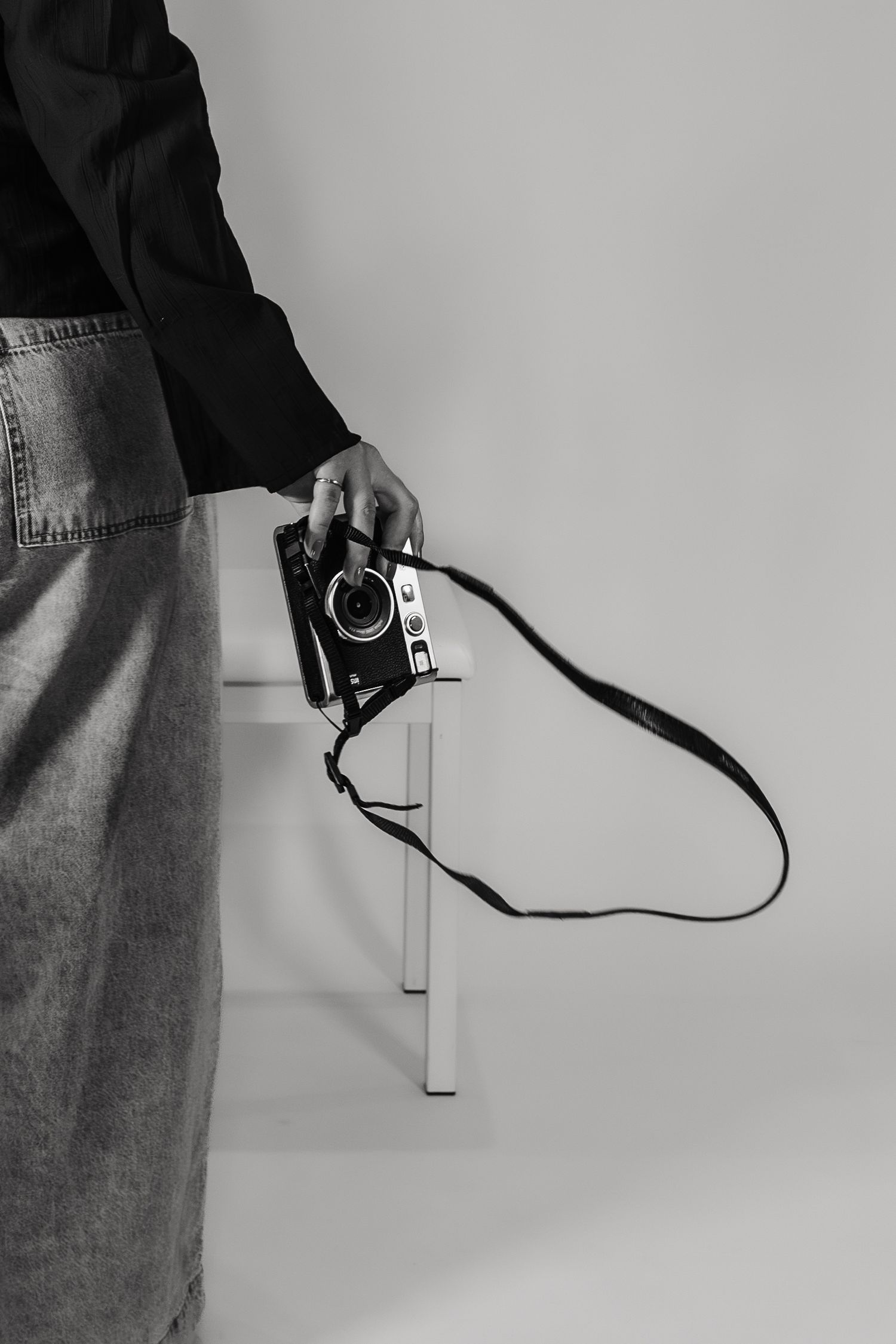 A hand holding a vintage camera with strap against white wall, wearing denim and dark clothing.