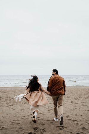A couple running together on a misty beach with waves in the background.