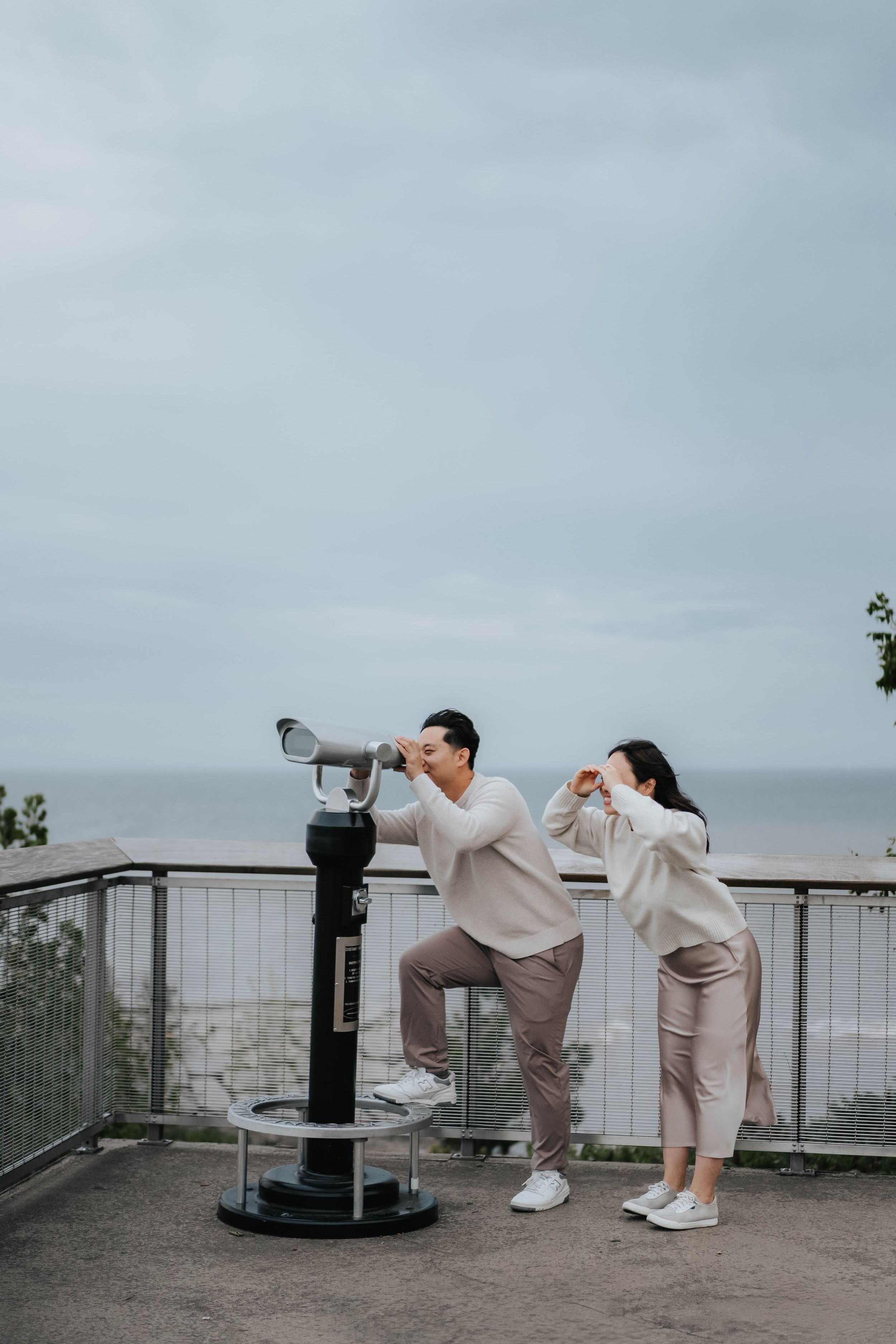 Two people in beige outfits look through viewfinder and binoculars at scenic overlook on cloudy day.