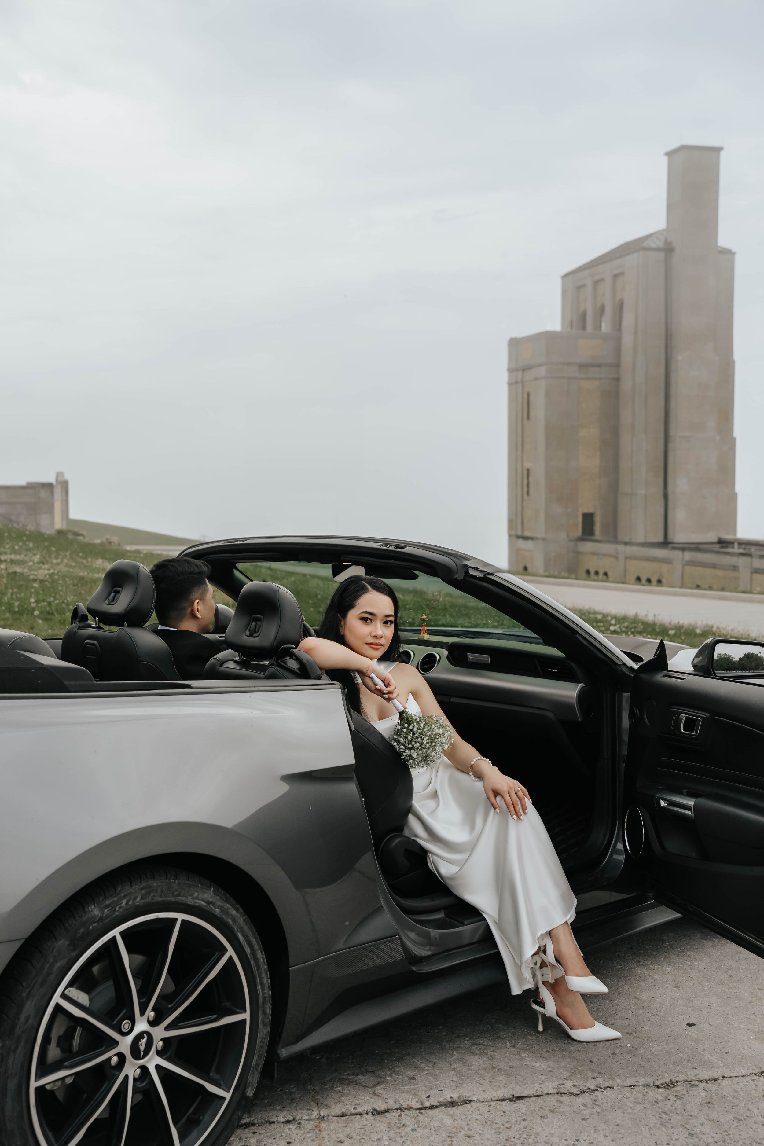 Elegant black car parked in front of industrial grain silos with person posing beside it.