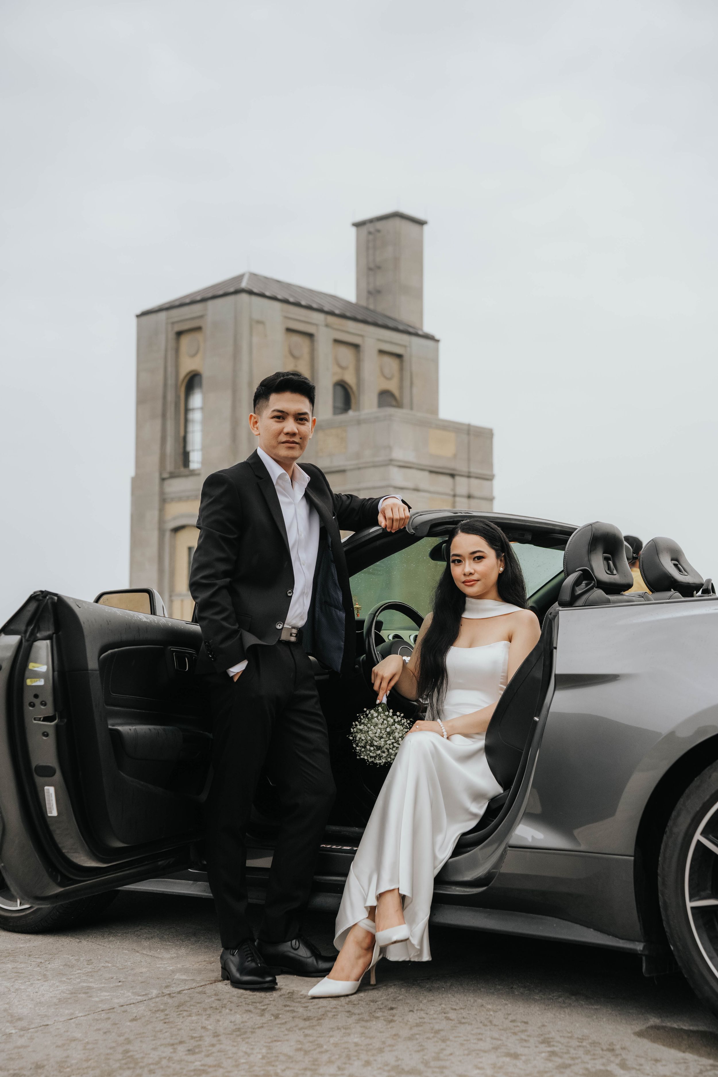 A couple in formal attire poses with a sleek black sports car in an urban rooftop setting.