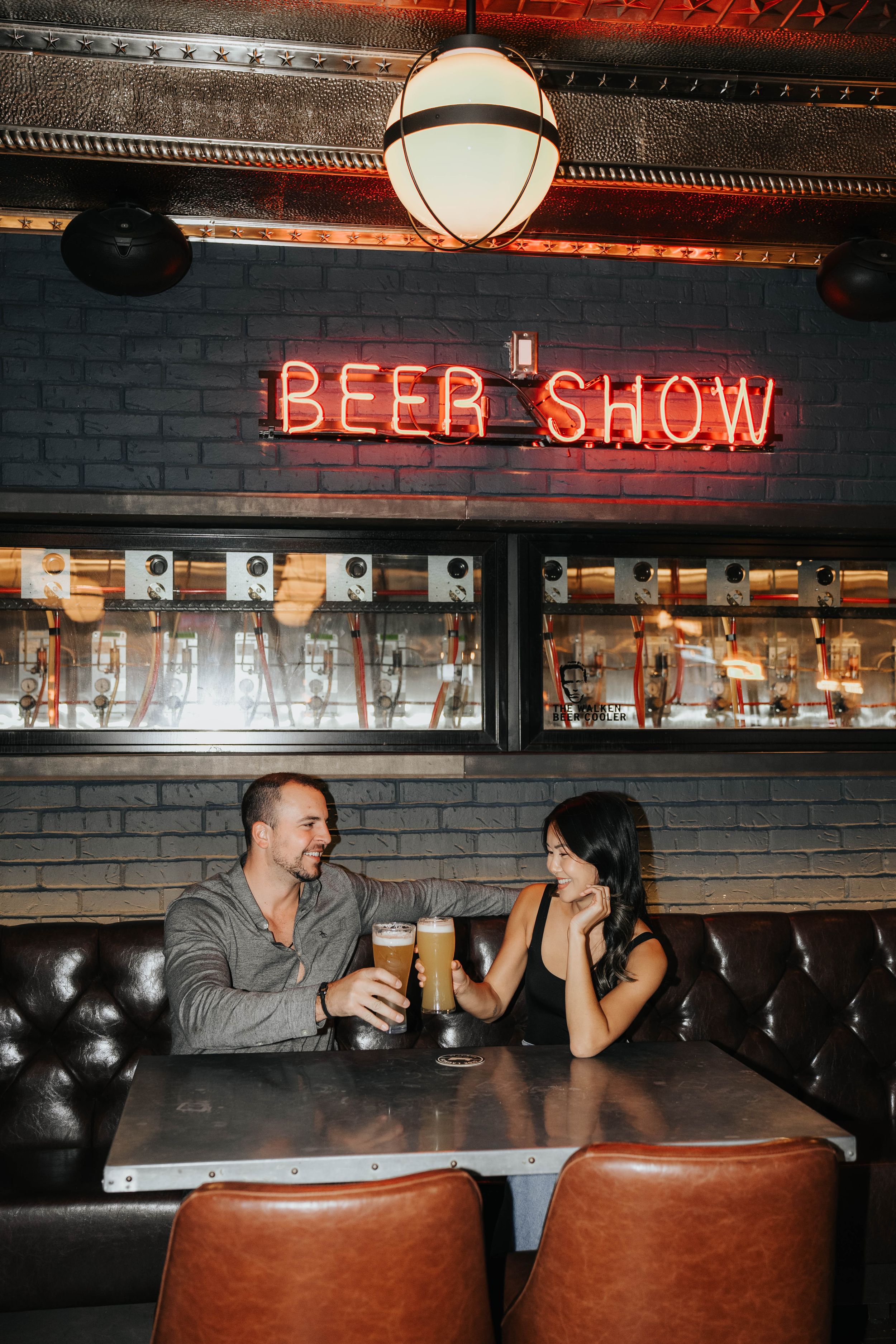 A couple enjoys drinks at a bar with a red neon 'BEER SHOW' sign and leather booth seating.