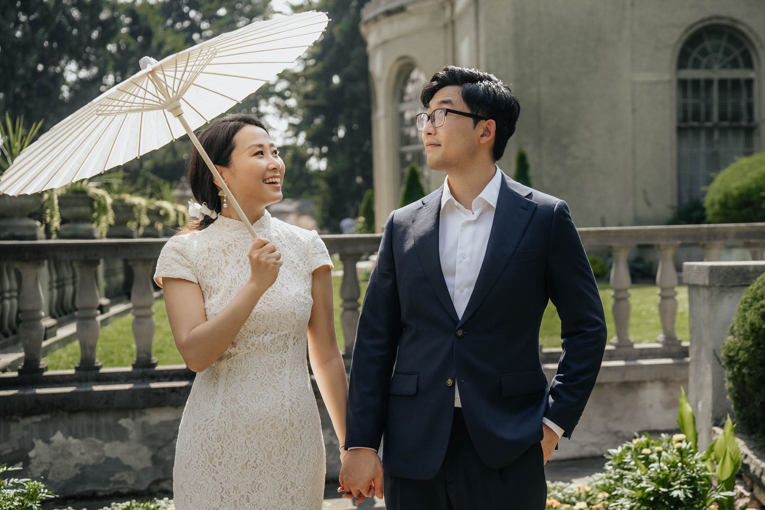 A bride and groom share romantic moments in a garden setting with a white parasol and elegant formal attire.