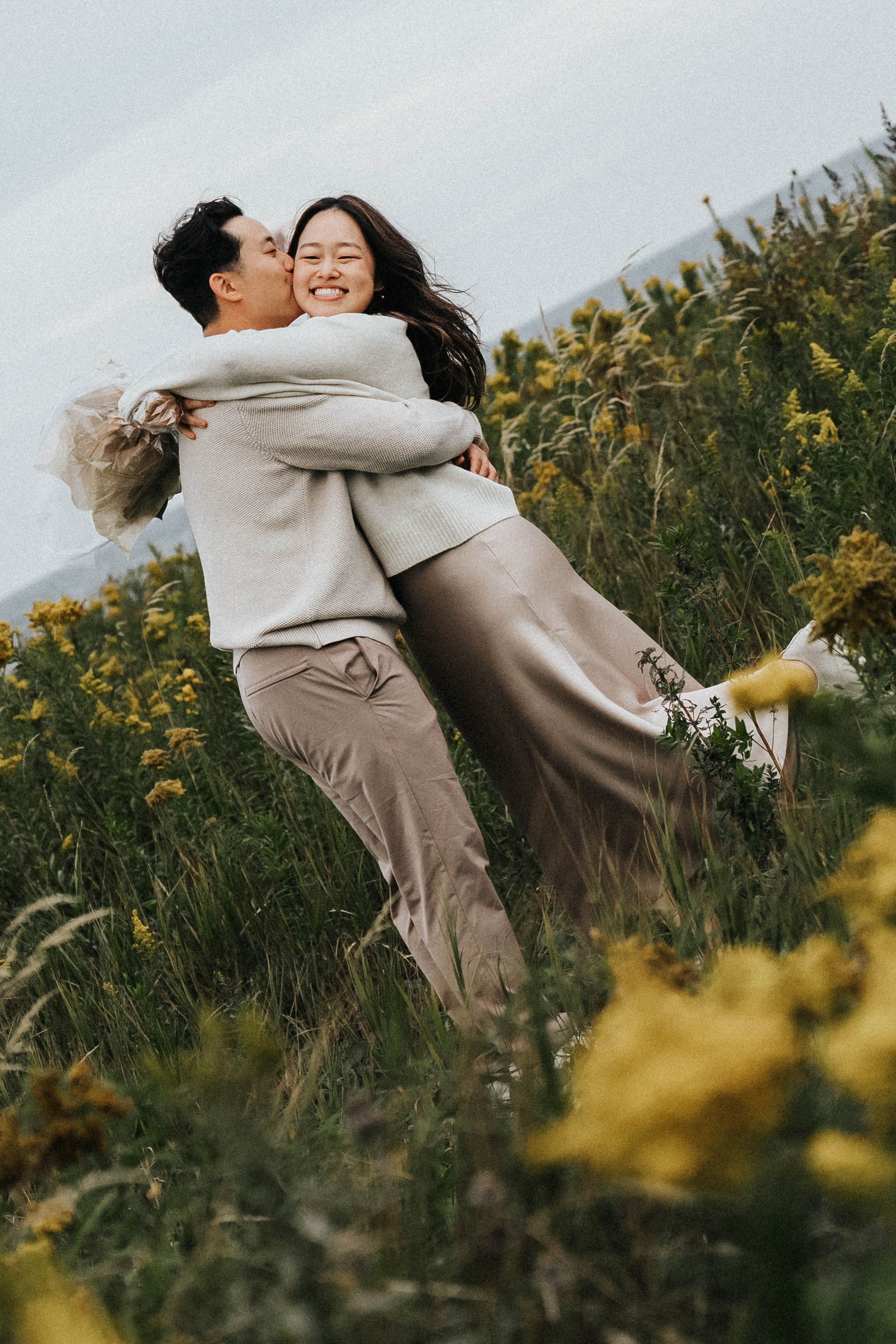 A couple embraces joyfully in a field of yellow wildflowers on a sunny afternoon.