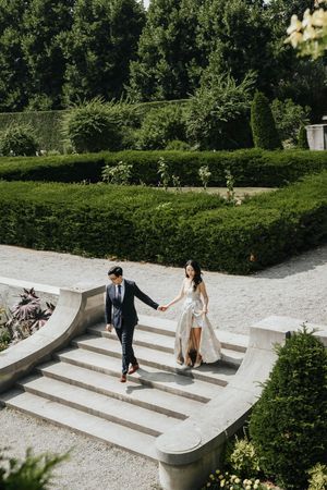 A couple descends elegant stone steps surrounded by lush greenery in a garden setting.