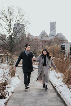 Two people in winter coats walk together on a snowy path while holding hands and smiling.