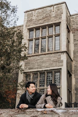 Two students sit and chat on a stone ledge outside a gothic style university building on an autumn day.
