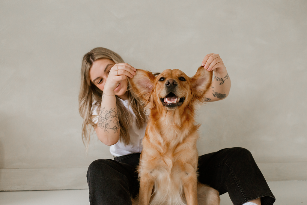 Girl sitting against a wall with her dogs ears held up and dog is smiling at the camera.