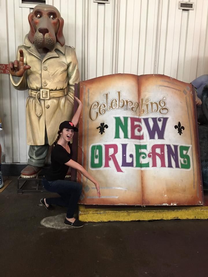 Girl squatting beside sign and holding hands up showcase in the sign that says "Celebrating New Orleans".