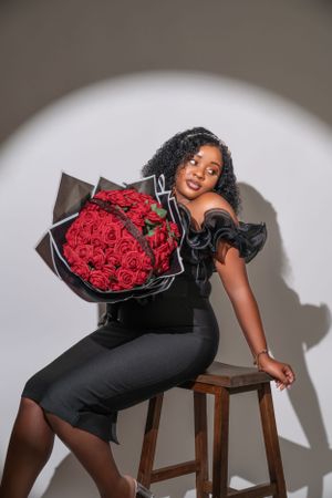 Elegant portrait against white background featuring black dress, wooden stool and dramatic bouquet of red roses.