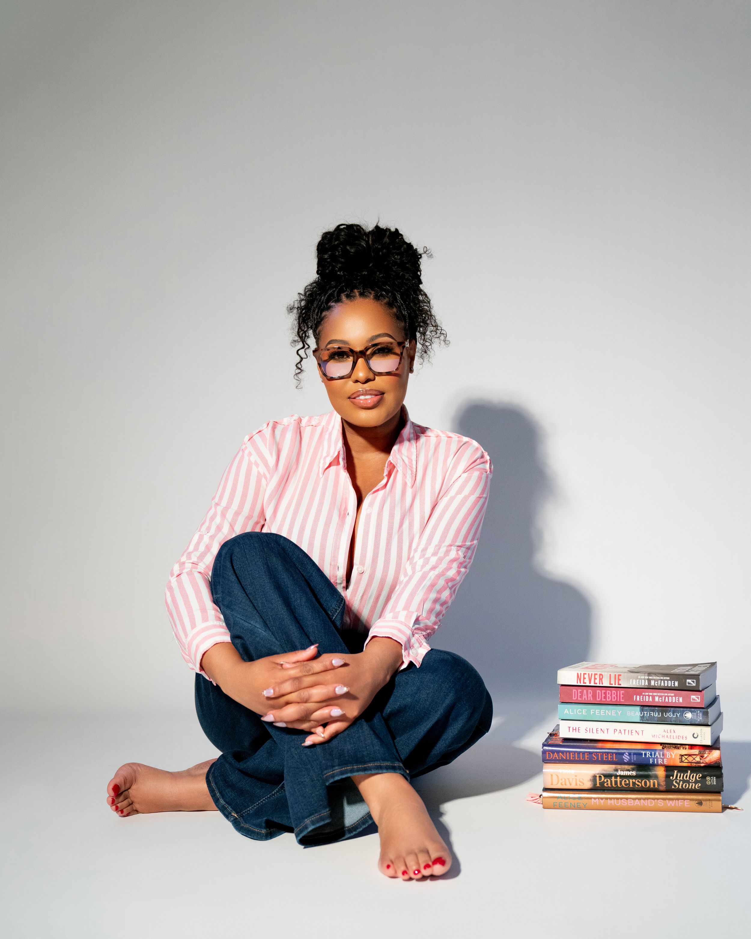 Woman with glasses and curly updo sits cross-legged beside a stack of books, wearing a pink striped shirt and jeans.
