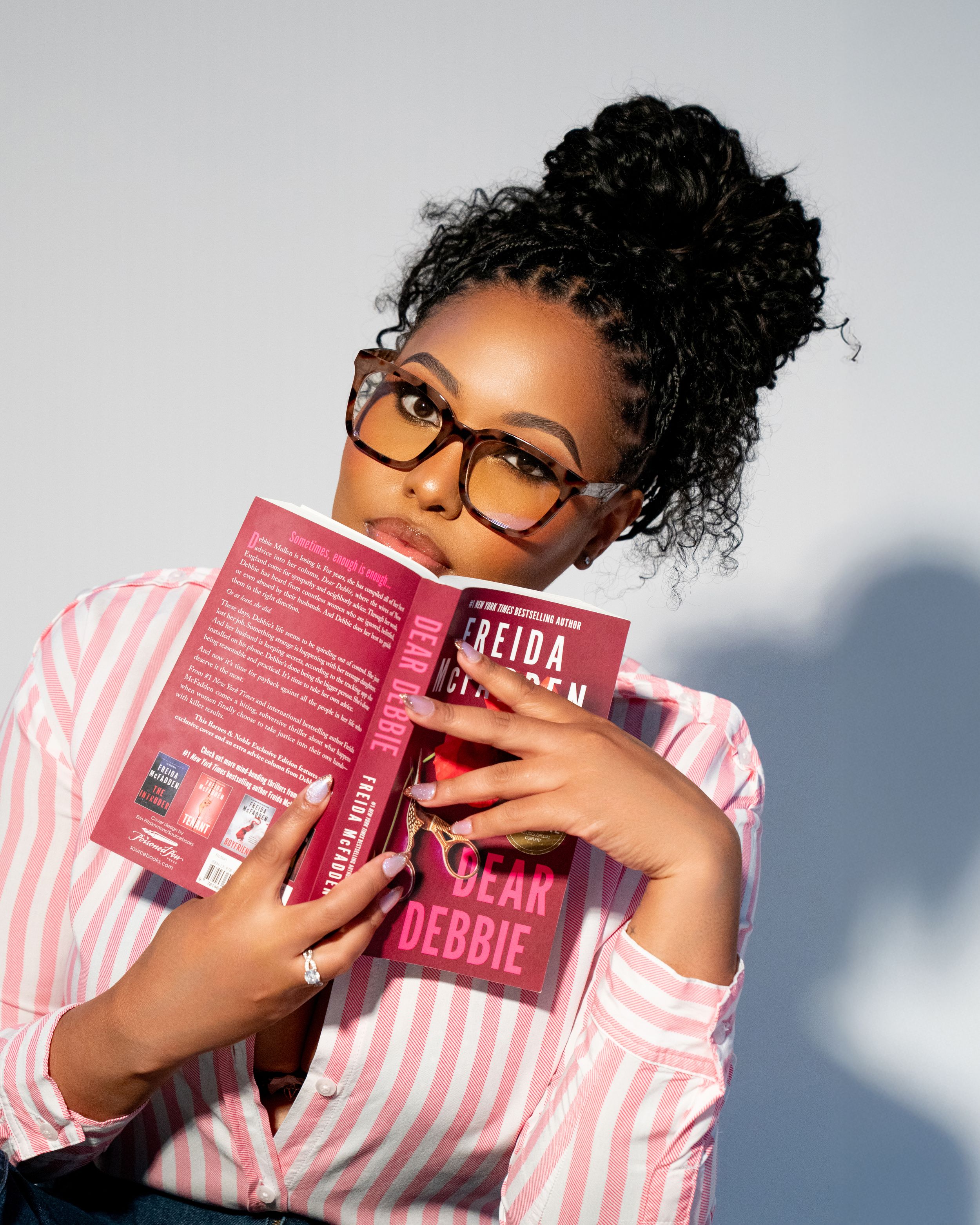 Young woman with glasses and curly updo holds a pink book titled Dear Debbie, wearing a striped shirt.