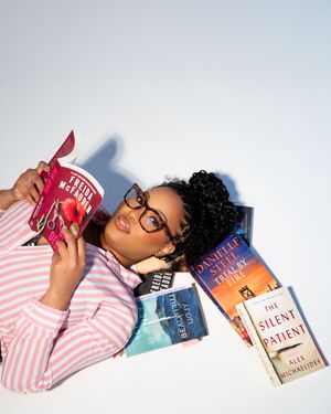 Woman lying on floor surrounded by books including The Silent Patient, wearing glasses and pink striped shirt, reading enthusiastically.