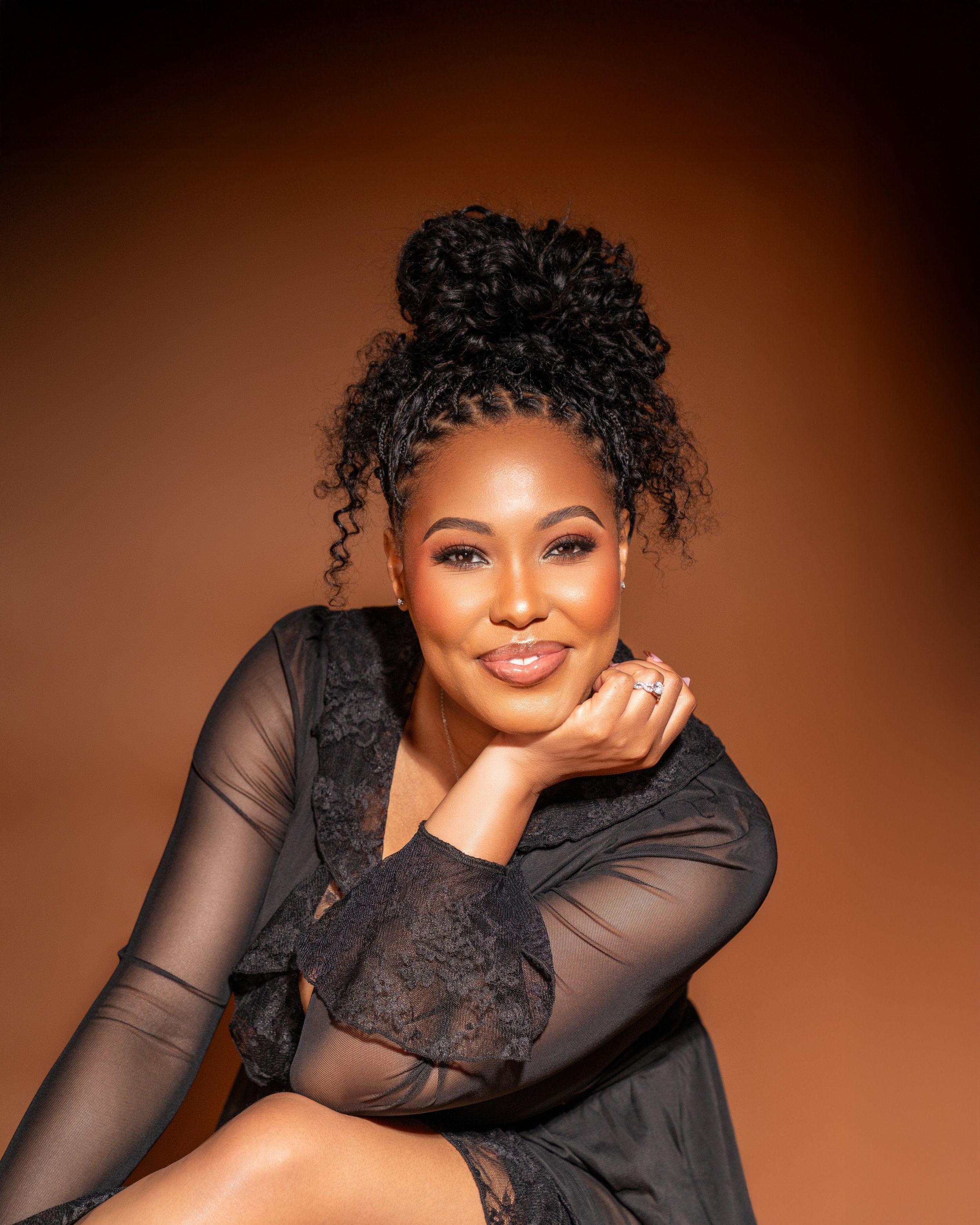 A confident Black woman in a black lace outfit poses gracefully against a warm brown studio background.