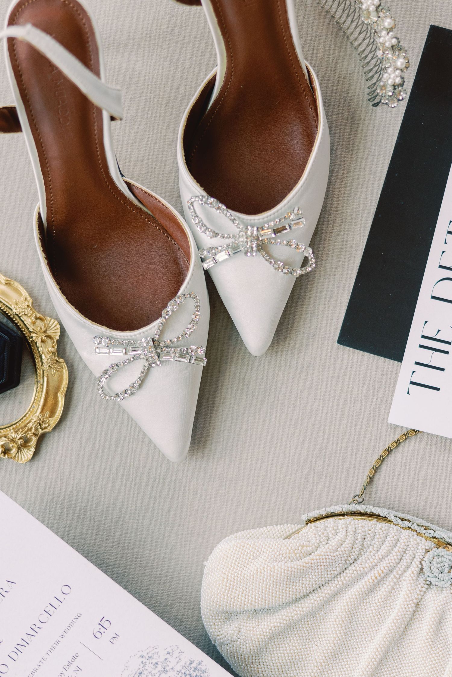 Elegant white bridal heels with crystal bow embellishments displayed alongside a pearl clutch and gold accessories.