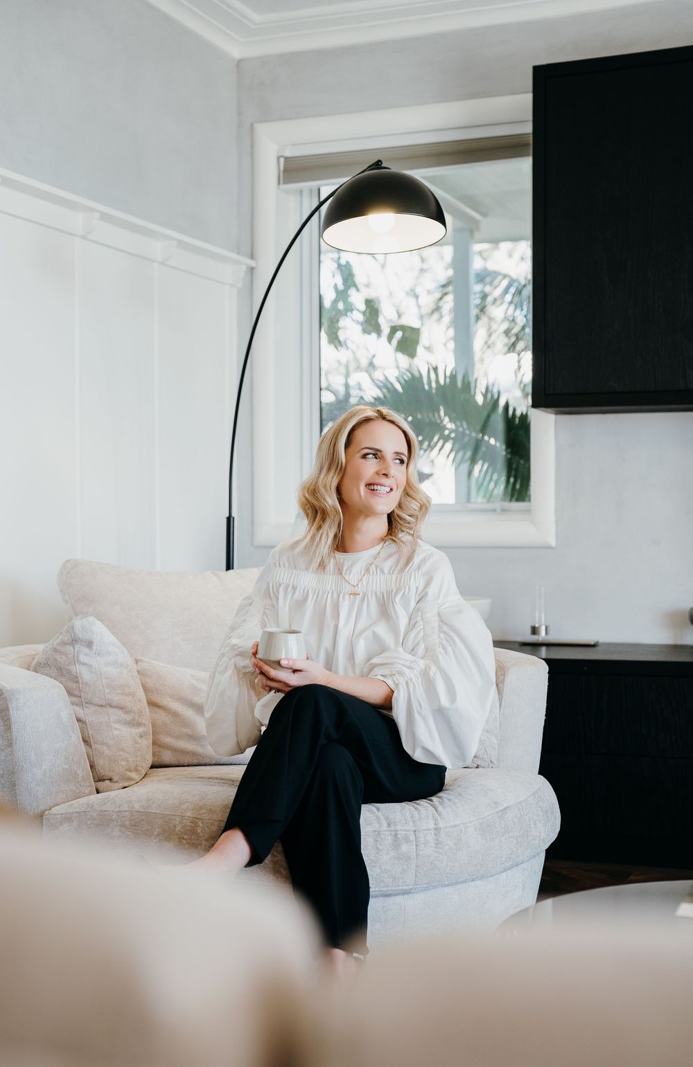 Woman relaxes on white couch in bright modern living room with black pendant lamp and window views.