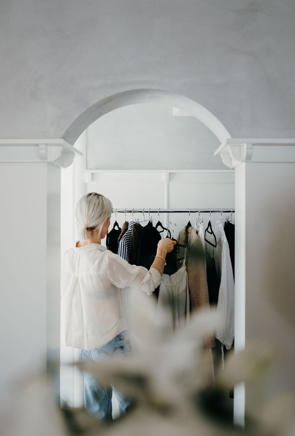 Person in white clothing organizing garments on clothing rack against white wall in minimalist space.