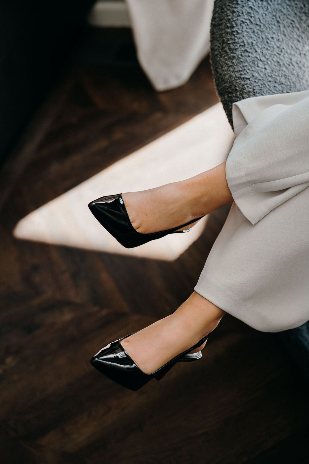 A close-up view of black patent leather pointed toe heels worn with a white dress against a dark wooden floor.