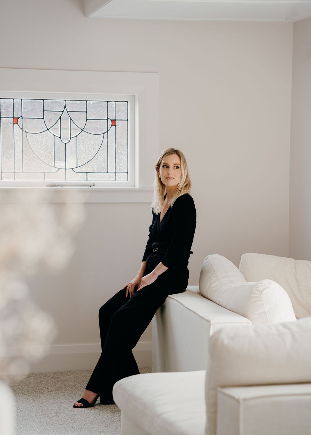 A figure in a black outfit sits and poses in different positions against white walls and furniture in a minimalist setting.