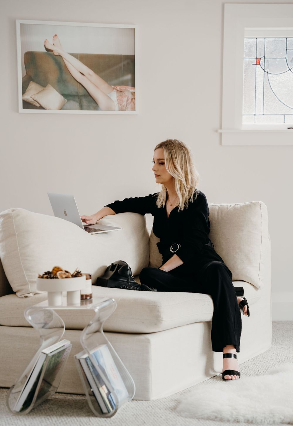 A person in black attire relaxes on a white sofa while working on a laptop in a minimalist room setting.