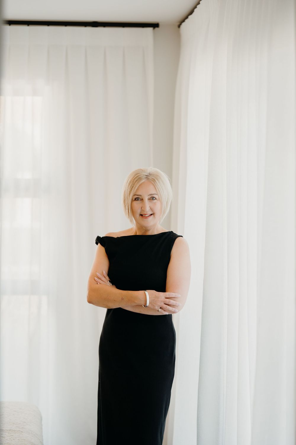 A woman in an elegant black sleeveless dress stands confidently with crossed arms against white curtains in minimalist setting.