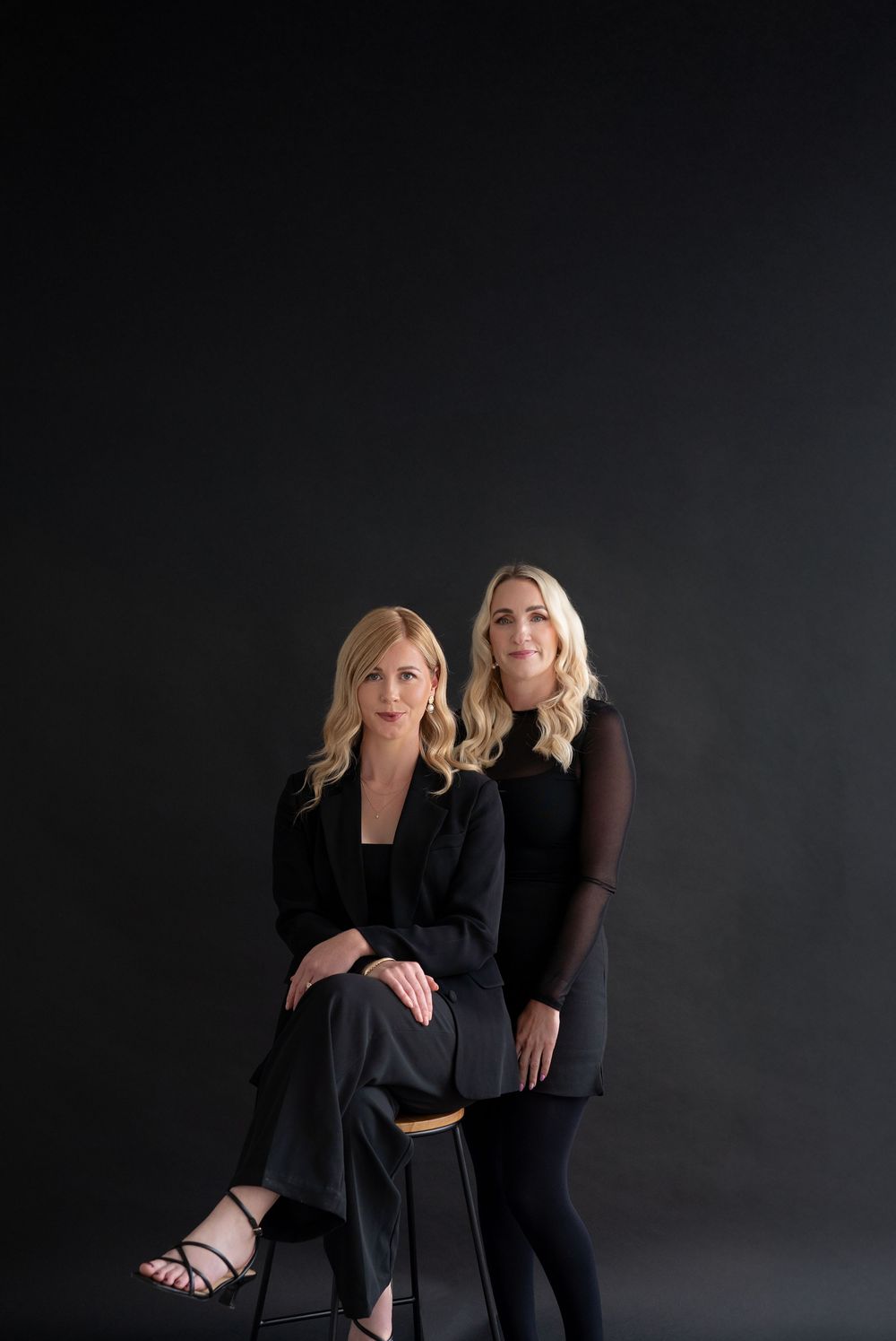 Two women in black outfits pose elegantly against a dark studio backdrop for a professional portrait.
