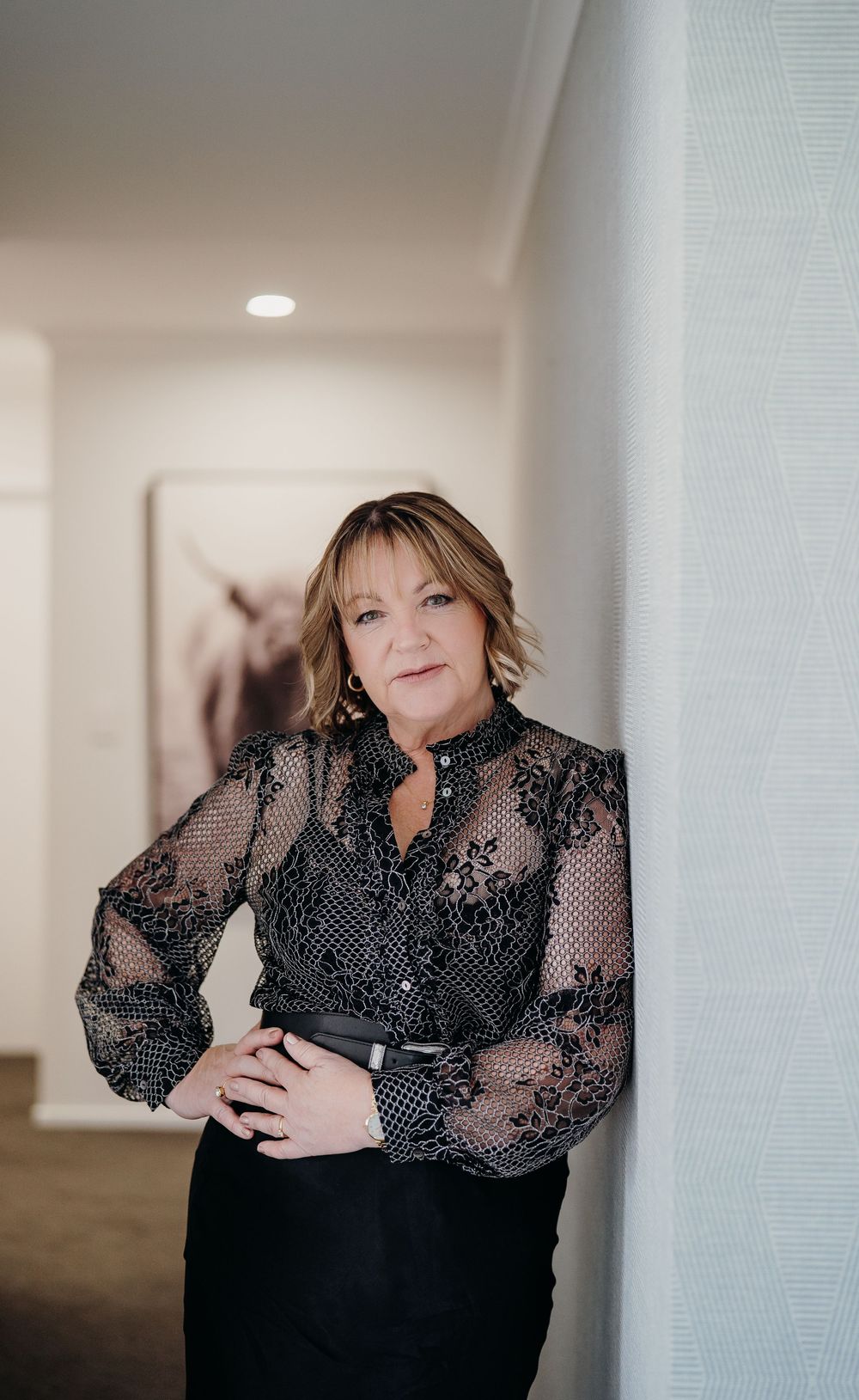 Woman in elegant black lace blouse with sheer sleeves poses against white wall in indoor setting.
