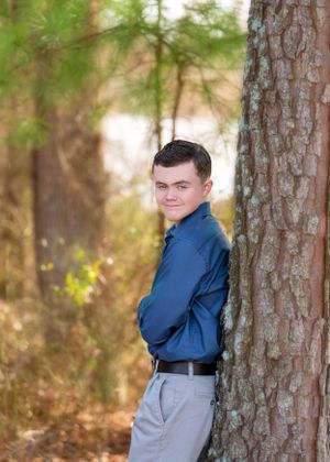Portrait of young man in blue shirt posed against large tree trunk.