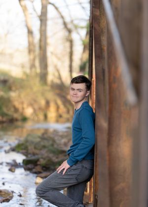 Senior portrait of young man in blue sweater leaning against rustic metal structure.