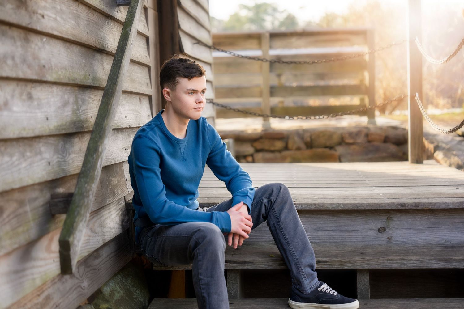 Young man in casual blue sweater sitting on wooden deck during outdoor photoshoot.