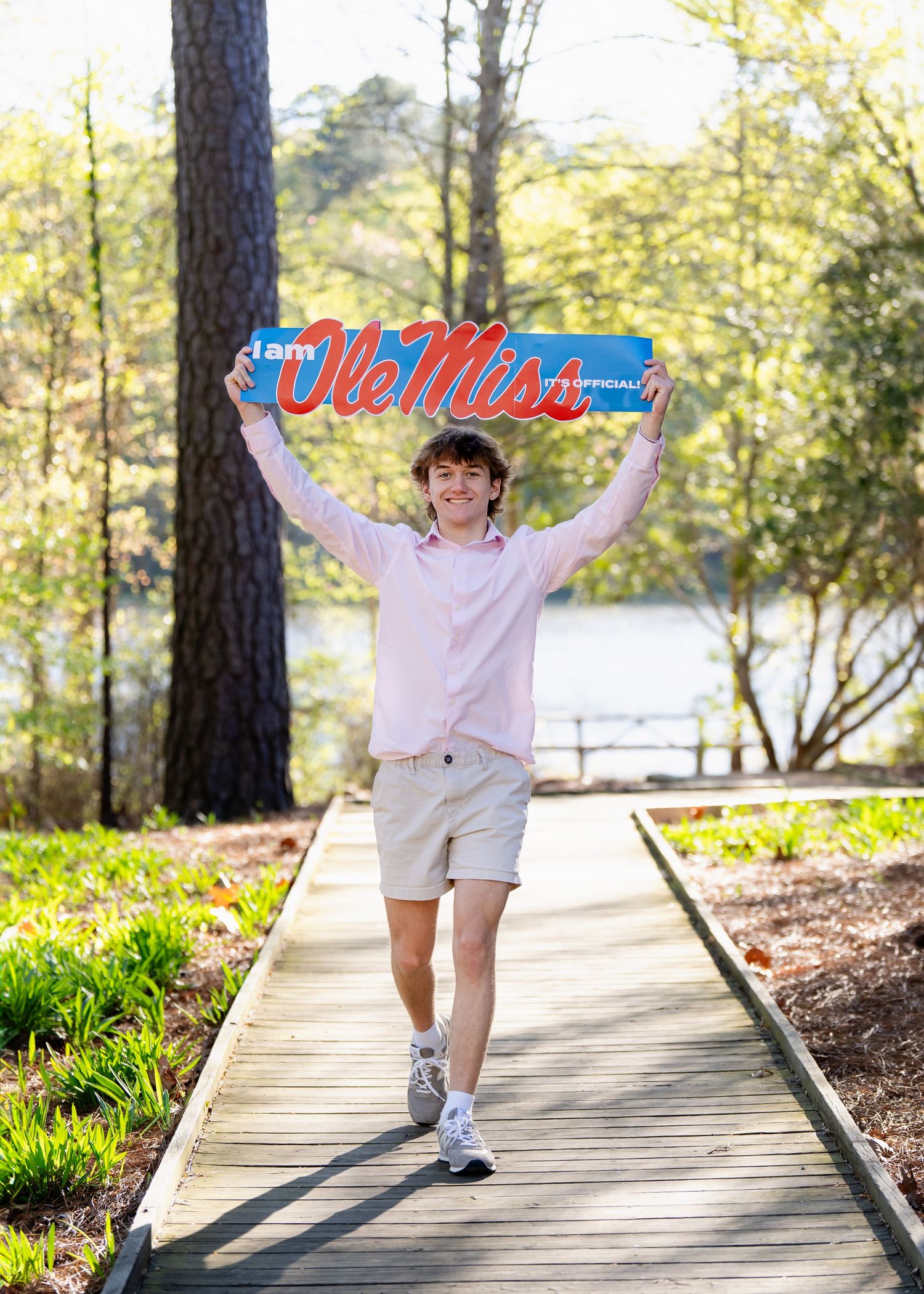 Student celebrating with Ole Miss banner on wooden boardwalk surrounded by trees.